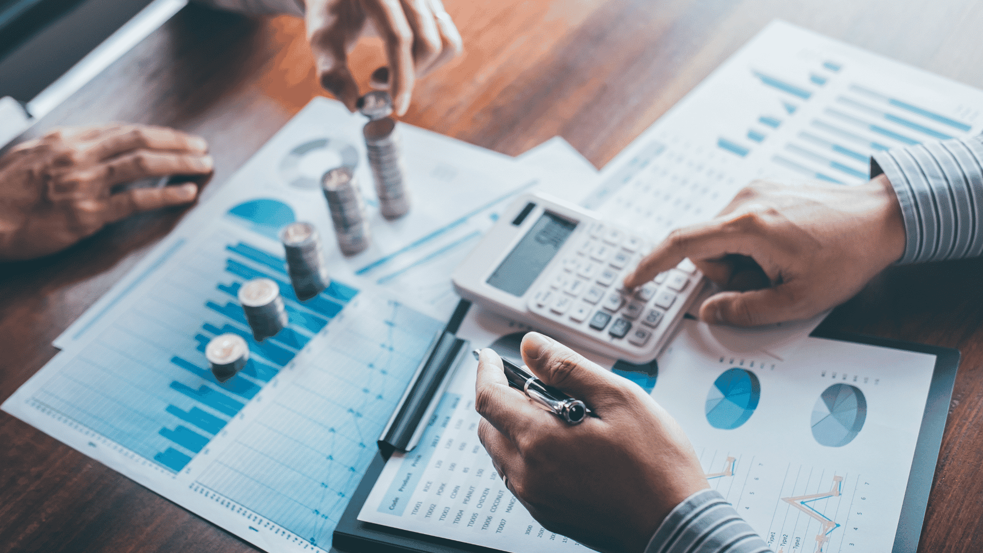 Mãos aparecendo calculando o financeiro de uma empresa, para representar tem uma calculadora, pilhas de moeda e folhas de papel com gráficos.