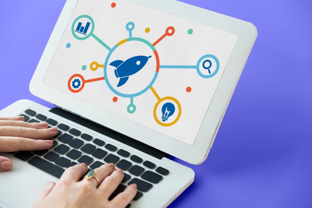 Uma mão usando um laptop com a tela mostrando ilustrações de foguete, engrenagem de configurações, gráfico, lupa e uma lâmpada, tudo representando a criação de um plano de ação 5w2h.