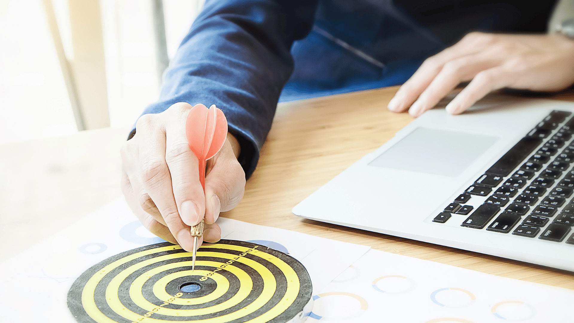 Uma mão masculina ao lado de um laptop e a outra mão cravando um dardo em um alvo que está em uma folha de papel, indicando a escolha do público-alvo.