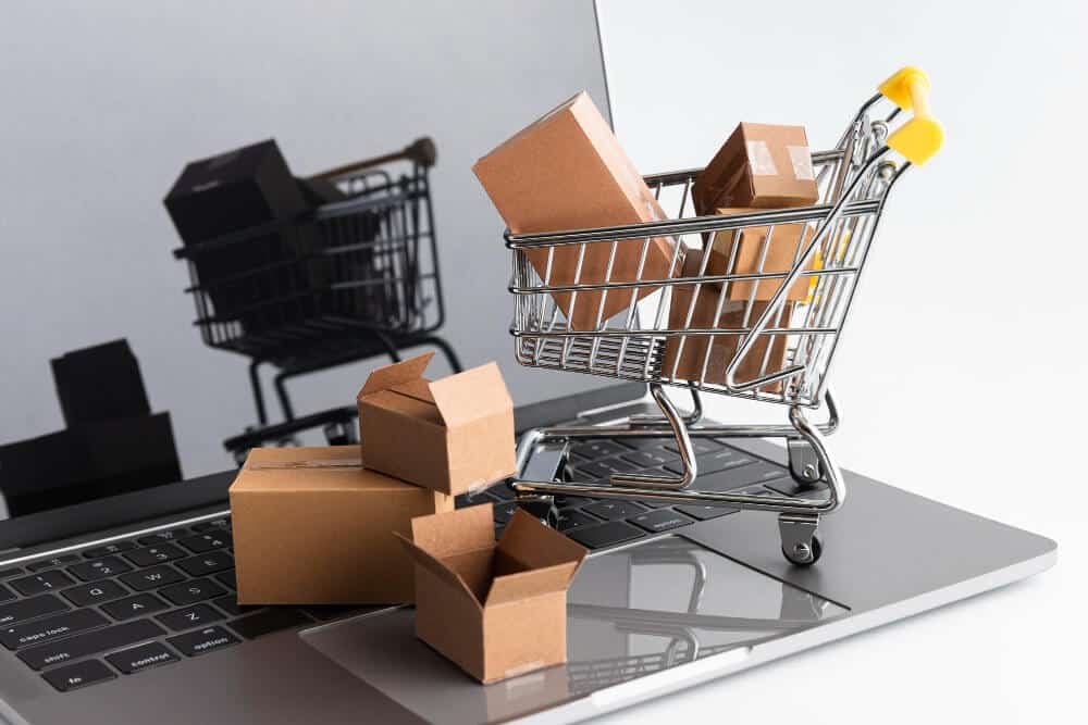 Imagem com um notebook aberto e em cima dele um carrinho de compras com caixas de papelão