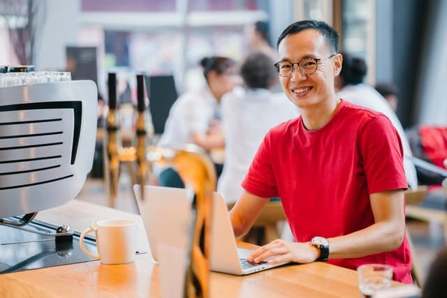 Homem de camiseta vermelha sorrindo utilizando um notebook, com outras pessoas ao fundo